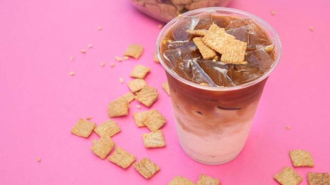 A bright pink background. A plastic cup full of milk layered with coffee and cinnamon toast crunch squares sprinkled across the background.