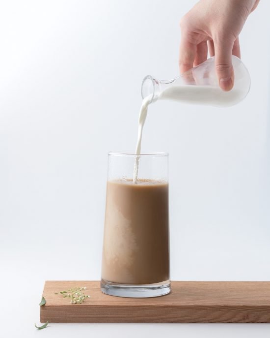 A glass full of coffee and milk sits on a counter. A singular hand is pouring an alternative milk into the same glass.