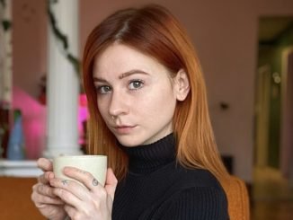 Casey is a young white woman with red hair. Casey sits on a rust orange couch with green plants and white columns in the background. She wears a black turtleneck with three-quarter length sleeves. She holds a white mug in her hand. She sits at a 45 degree angle, with her eyes angled toward the camera and her hair tucked behind her left ear.