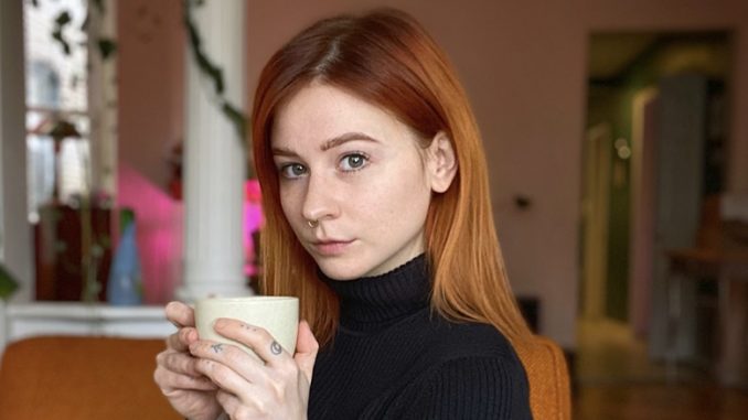 Casey is a young white woman with red hair. Casey sits on a rust orange couch with green plants and white columns in the background. She wears a black turtleneck with three-quarter length sleeves. She holds a white mug in her hand. She sits at a 45 degree angle, with her eyes angled toward the camera and her hair tucked behind her left ear.