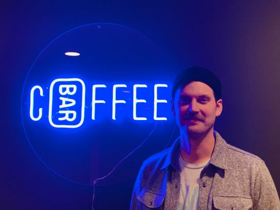 This is a picture of Kyle Anderson the founder of Coffee Bar Seattle. He is a white man wearing a black beanie and has a brown mustache. Behind him is a neon blue sign that says Coffee Bar.