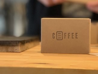 This is a picture of a soap bar made with coffee grounds. It is sitting on a wooden counter wrapped in cardboard packaging.