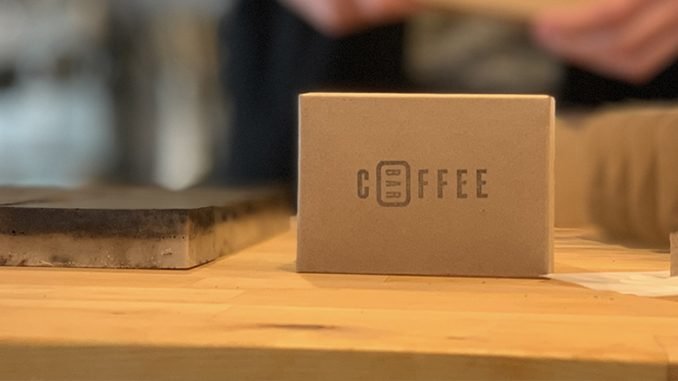 This is a picture of a soap bar made with coffee grounds. It is sitting on a wooden counter wrapped in cardboard packaging.