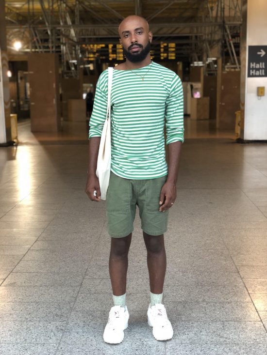 Adam is a bald, Black man with a beard. He stands in an industrial-looking building with gray, tiled floors and metal scaffolding along the ceiling. He wears a green and white striped shirt, green shorts, white sneakers, and carries an off-white tote bag on his shoulder. He looks directly into the camera with a neutral expression.
