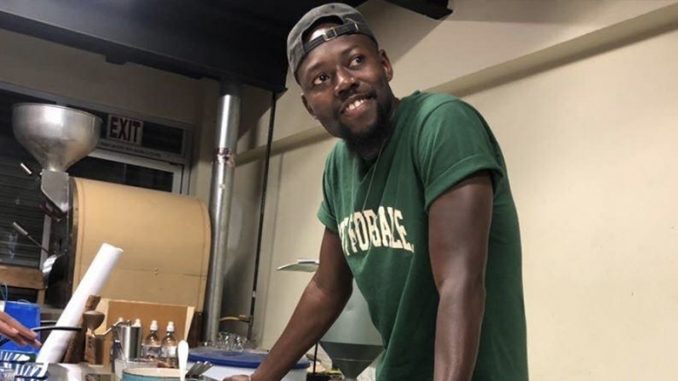 Baidy stands at a coffee cupping table smiling. He wears a bright green tshirt,