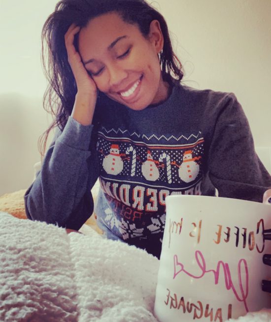 Court, a Black woman with medium-length, wavy brown hair, sits among white blankets while holding a coffee mug that says “coffee is my life.” She has a toothy grin, and her eyes appear closed. She wears a dark grey Christmas sweater with images of snowmen and candy canes.