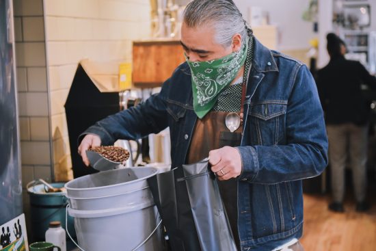 Paul is scooping coffee into a bucket. He is wearing an apron with his cupping spoon in it.