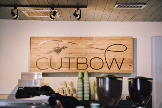 A shot of the cafe sign that read cutbow. We see the top of an espresso hopper and several to-go cup stacks in the back.