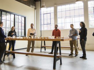 A group of people stand together inside a warehouse building. They are sampling coffee.