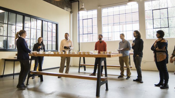 A group of people stand together inside a warehouse building. They are sampling coffee.