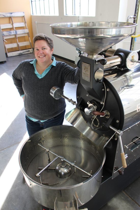 Joey Gleason is a woman with short cropped hair. She stands next to a large roasting machine in the Buckman Coffee Factory.