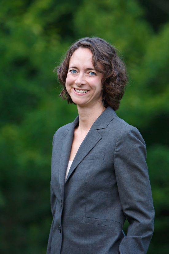 Kellem is a white woman in her middle age. She has short curly brown hair and smiles standing for a portrait. She wears a grey suit jacket amidst a green forest background.