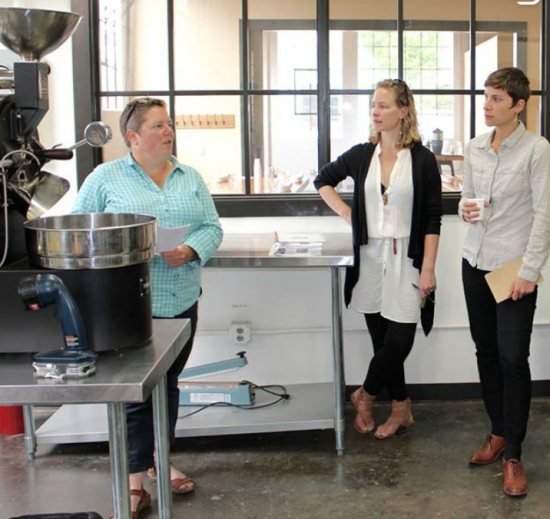 Joey stands next to the roaster. She is talking to two other individuals in the warehouse.