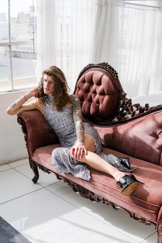 Veronica has long, brown hair and her right arm is covered in black tattoos. She wears a long, cold-shoulder silver dress with a slit down the side. Her leg is exposed from the thigh down, and she wears laced black heels. Her body is angled away from the camera, and she sits on an ornate, rust red couch.