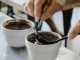 A close up of hands scooping coffee grounds out of a cup with two spoons. There are several coffee cupping samples around the silver table.