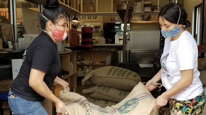 Two people stand in a coffee roastery, a small silver roaster visible in the background. They are lifting a brown burlap coffee bag onto a wooden pallet, presumably in preparation to be roasted.