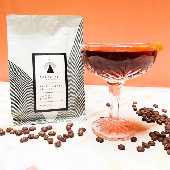 A coffee bag from Broadcast roasters sitting next to a cocktail glass full of liquid. It is on an orange and white background, and coffee beans are scattered on the table it is sitting on.