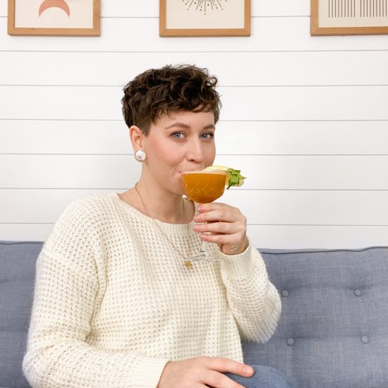 Molly is a young white woman. She sips on a non-alcoholic coffee coocktail. She has short brown hair and is wearing a white knitted sweater and sitting on a blue couch.