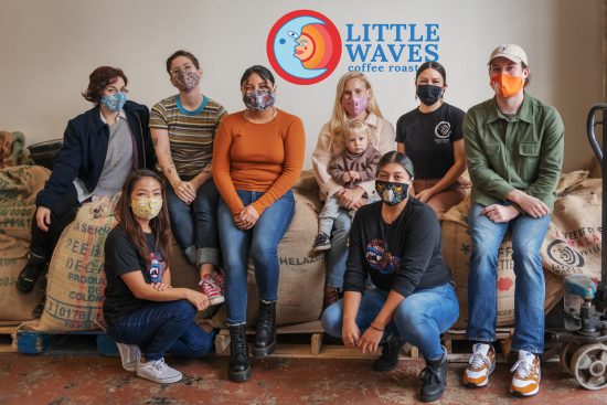 The Little Waves team, all wearing face masks, sits on top and in front of a number of large, burlap coffee bags. The Little Waves Logo is in the background, which features a blue crescent moon in front of a bright red and orange sun. The words “Little Waves Coffee Roasters” appear in blue lettering on the right of the logo.