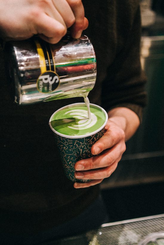 A closeup shot of hands pouring latte art into a hot cup filled with matcha latte.