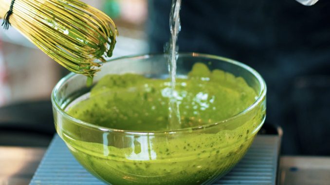 A closeup of a clear bowl filled with matcha. Above it is a bamboo whisk.