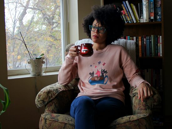 Niki sits on a floral armchair, with their arm perched up on one side, and the other hand holding a red coffee cup that features an image of a mouth. She wears turquoise, square-rimmed glasses, jeans, and a pink sweater with an image of cats throwing glitter out of a latte cap. She sits in front of a well-stocked bookcase.