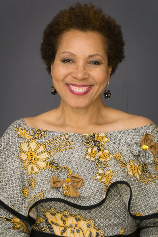 Phyllis has short, brown hair and wears ornate, black and gold earrings. Her blouse is flowy, ornate, and features gold and brown flowers on a black, checkered background. Image features Phyllis from the stomach upwards.