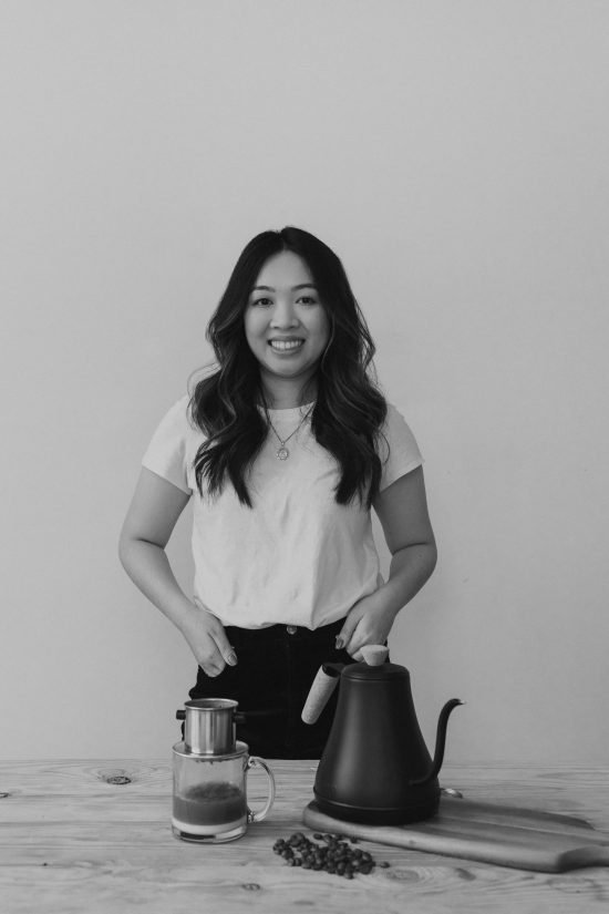 Kim is a young woman with long curled hair. She wears a white t shirt and smiles in front of a coffee brewing set.e