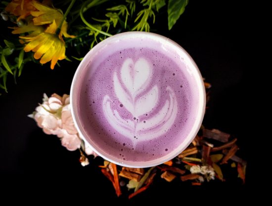 A paper coffee cup with a purple drink in it. There is a rosetta. The cup sits on a bed of flowers.