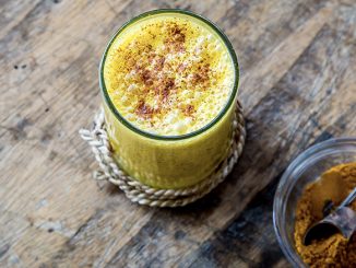 There is a bright yellow turmeric milk in a glass centered. Next to it is a mason jar filled with turmeric powder.