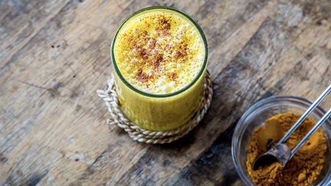There is a bright yellow turmeric milk in a glass centered. Next to it is a mason jar filled with turmeric powder.