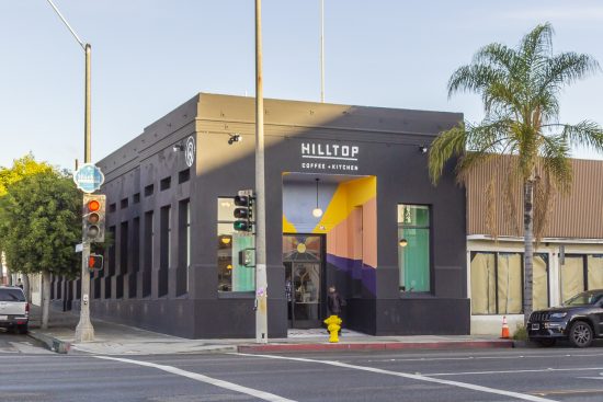 The storefront of hilltop in Inglewood pictured. It is a square shaped black building with plain white font saying hilltop. It is a bright and sunny day and a palm tree stands out front of the cafe.