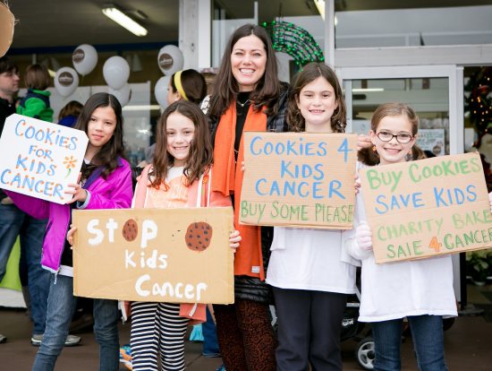Gretchen is the founder of Cookies for CAncer, standing and smiling with a group of child volunteers. She is a middle age white woman with long brown hair.