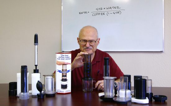 AeroPress founder Alan Adler in a man in his later ages sitting at a desk posing. He presses the aeropress, and several pieces of it lie on the table in front of him.