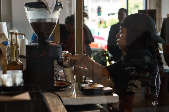 A barista pulls a shot of espresso at the cafe. She wears a black beanie and has dreads.