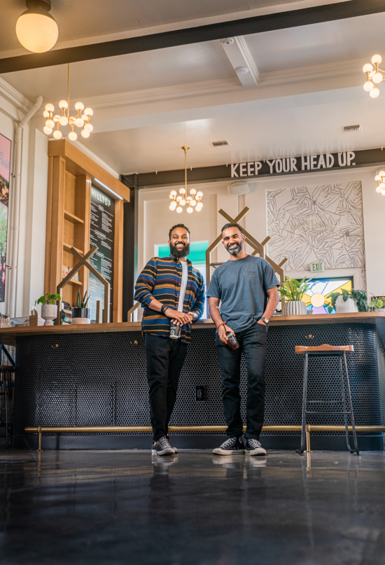 Yonnie and Ajay stand smiling with their hands in their pockets inside one of the hilltop locations. Ajay wears a multicolor collared shirt and Yonnie has a plain grey t shirt.