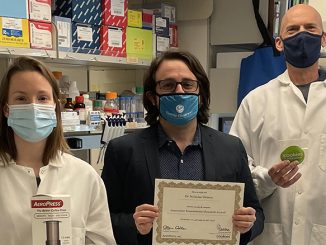 Dr. Vitanza and his lab founders stand with face masks posing for a photo. To the left, a Carry Myers holds an AeroPress. Dr. Vitanza is in the middle holding the grant paper, and to the right is Matt Biery holding stickers that say Cookies for Cancer.