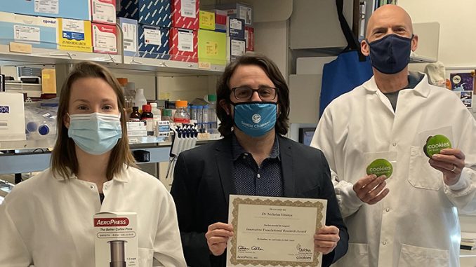 Dr. Vitanza and his lab founders stand with face masks posing for a photo. To the left, a Carry Myers holds an AeroPress. Dr. Vitanza is in the middle holding the grant paper, and to the right is Matt Biery holding stickers that say Cookies for Cancer.