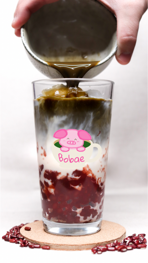 Closeup of a large glass with a Bobae pig cartoon logo. A hand pours a beverage into the glass filled with boba pearls.