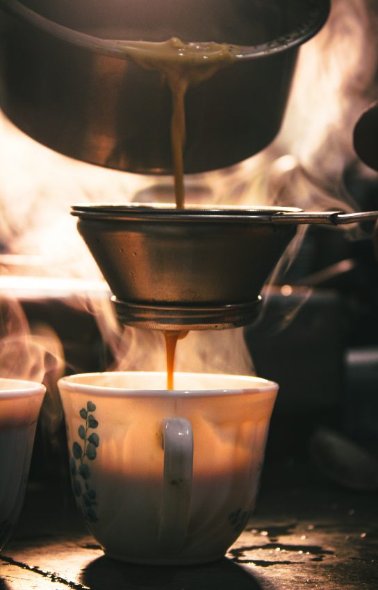 A closeup of a metal tea steeper. Above it liquid is being poured through, and a mug sits underneath the tea steeper with chai.