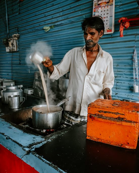 A man in India pours hot milk between a pot and a pitcher to cool and froth it. This happens in a commercial setting with a blue kitchen.