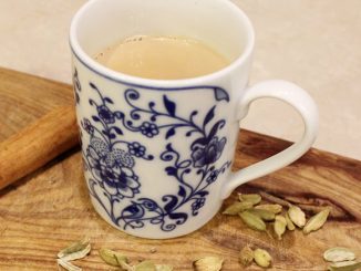 A colorful mug full of light brown chai sits inside. The front of the mug has blue flowers and it sits on a wooden table with cardamom seeds and a cinnamon stick.