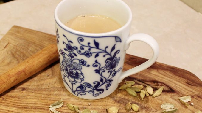 A colorful mug full of light brown chai sits inside. The front of the mug has blue flowers and it sits on a wooden table with cardamom seeds and a cinnamon stick.