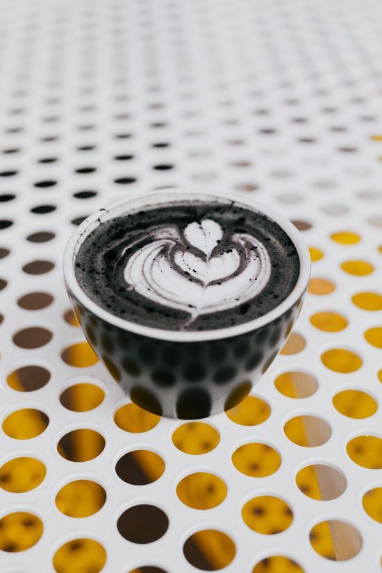 A polka dot background of a table with a cup on it. In it is a charcoal latte with a black and white rosetta drawn into it.