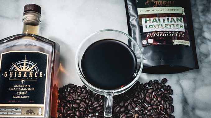 A closeup of a glass mug holding brewed coffee. Underneath it are coffee beans, and on either side of the mug rest the bag of Haitian coffee beans and the bottle of whiskey.