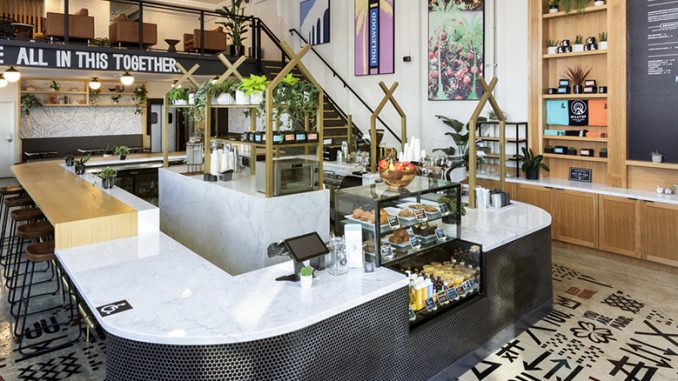 The interior of a hilltop cafe location. It has tall ceilings, chandeliers, and a mosaic tiled floor. The bar front is marble top and house plants hang amidst the backdrop.