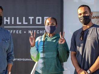 Ajay, Issa, and Yonnie stand pictured in front of one of the Hilltop locations. On the left, Ajay had short cropped hair and a blue chambray shirt. Issa is in the middle pictured with a singular long braid and a hoodie. Yonnie to the right has a plain grey shirt on with his hands crossed. All are wearing face masks.