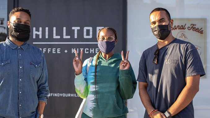 Ajay, Issa, and Yonnie stand pictured in front of one of the Hilltop locations. On the left, Ajay had short cropped hair and a blue chambray shirt. Issa is in the middle pictured with a singular long braid and a hoodie. Yonnie to the right has a plain grey shirt on with his hands crossed. All are wearing face masks.