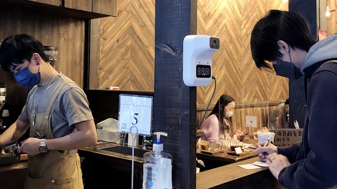 A barista rings up a coffee drink order to the left. To the right, a customer fills out a paper contact tracing application. They are both wearing masks and the scene is dimly lit.