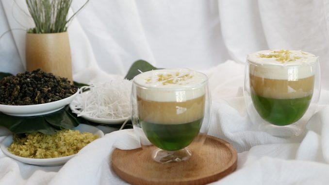 A set of pandan desserts sit on trays with a white sheet as a background. They are cups holding an array of tan, white, and green gelatins.